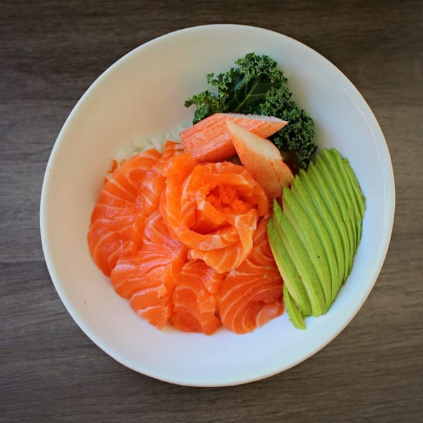 A white bowl filled with salmon sashimi, avocado slices, imitation crab, and kale.