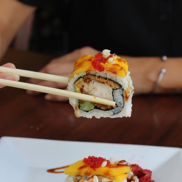 Chopsticks holding a Thai West sushi roll with mango, red roe, and white toppings.